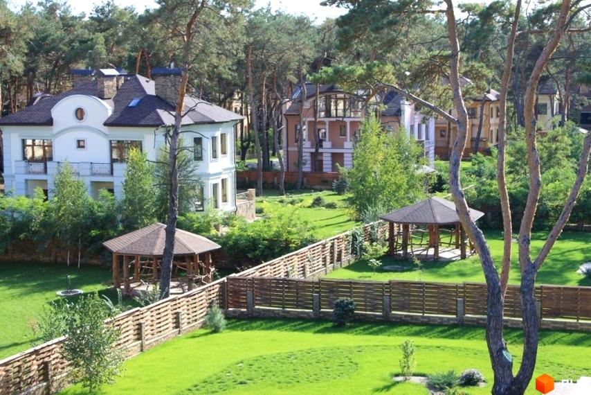 Двоповерховий будинок з білими стінами та темним дахом у Котеджному містечку Шишкино. На території є альтанки, доглянутий газон та дерев'яний паркан.