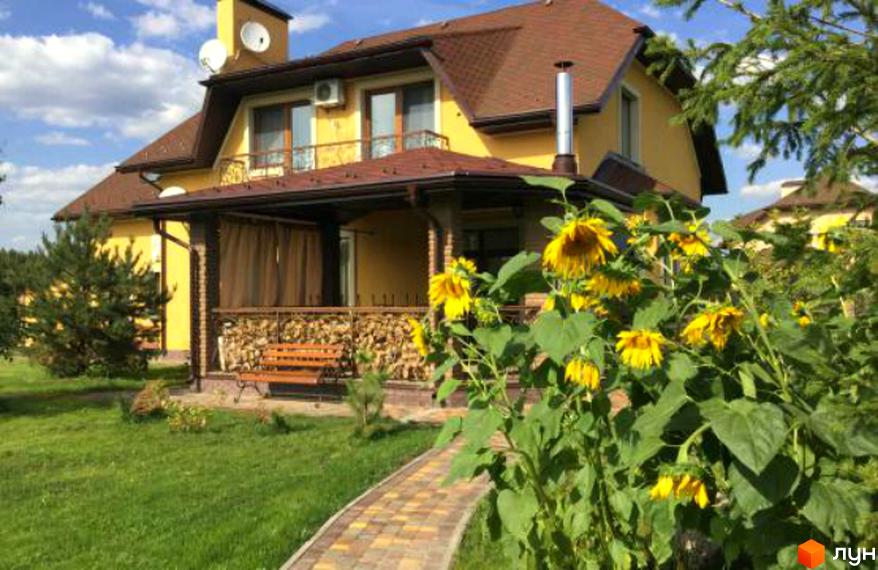 Житловий комплекс Swiss Village з жовтими стінами та коричневим дахом. Перед будинком доглянута прибудинкова територія з доріжкою та соняшниками.
