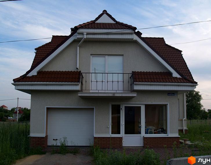 Будинок з коричневою черепичною покрівлею, балконом та гаражем. Навколо будинку зелена трава та рослинність.