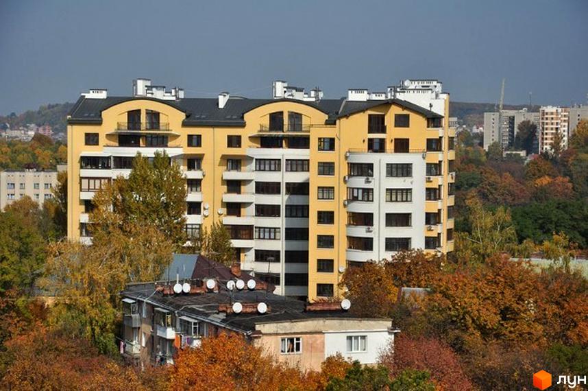 Житловий комплекс на вул. Героїв Крут, 11б має жовто-білі фасади та темні дахи. Будинок оточений деревами з осіннім листям та іншими будівлями на фоні.