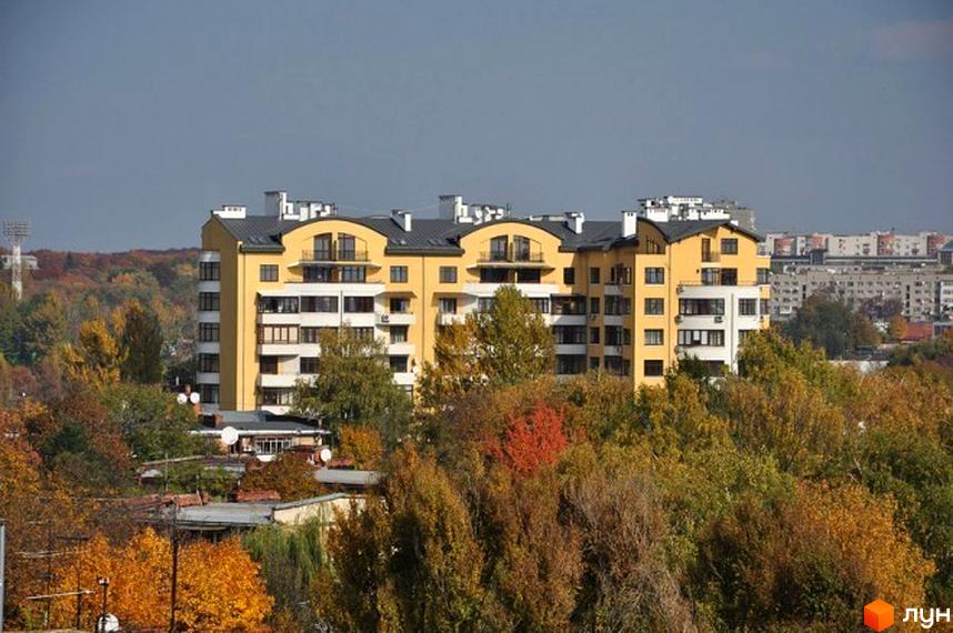 Житловий комплекс на вул. Героїв Крут, 11б, має жовті та білі фасади з темними дахами. Будівля оточена осінніми деревами та міським пейзажем.