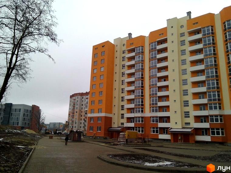 Багатоповерховий житловий будинок з помаранчевими та світло-жовтими фасадами, численними вікнами та балконами. Прибудинкова територія з вимощеною доріжкою.