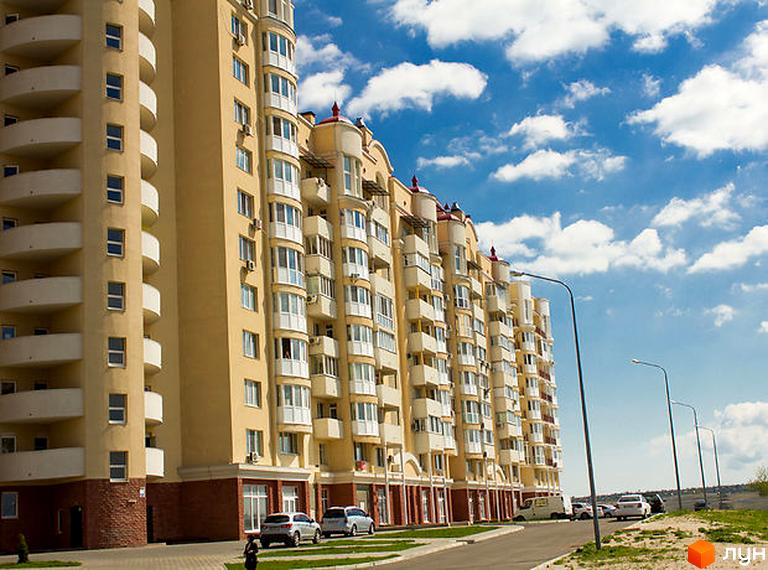 Житловий комплекс Рів'єра - фасади будівель з балконами та вікнами на тлі блакитного неба з хмарами. Поруч дорога та припарковані автомобілі.