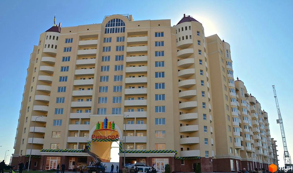 Зовнішній вигляд новобудови ЖК Рів'єра з численними балконами та вікнами під яскравим сонячним промінням. На фасаді будівлі видно вивіску з назвою комплексу та декоративні елементи.