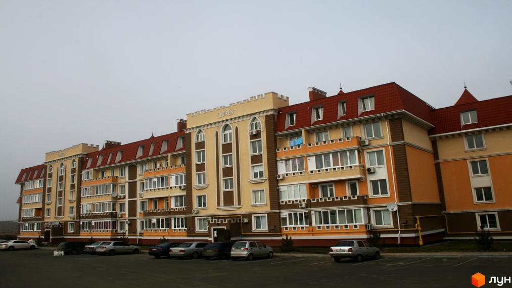 Зовнішній вигляд новобудови ЖК Академгородок-Совиньон з червоним дахом та бежево-коричневими фасадами. Перед будинком розташована парковка з автомобілями.
