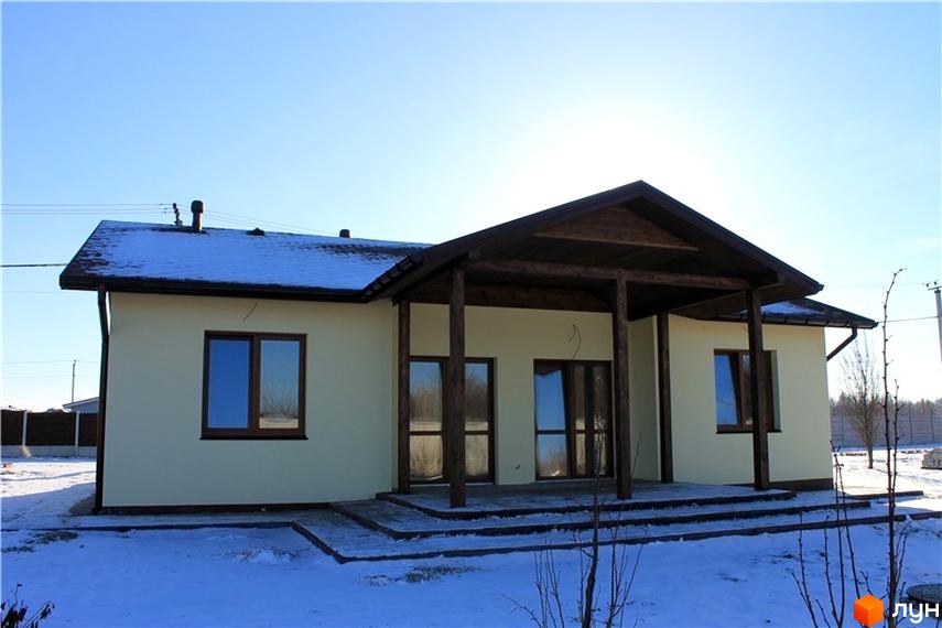 Котеджне містечко Рябинки: одноповерховий будинок з коричневим дахом та дерев'яною верандою на тлі блакитного неба та засніженої ділянки. Зовнішній вигляд будинку та прибудинкової території.