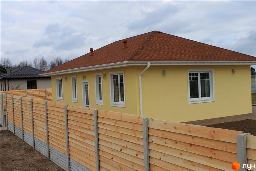 Котеджне містечко Рябинки: одноповерховий жовтий будинок із коричневим дахом та дерев'яним парканом. Навколо будинку видно зелені насадження та елементи ландшафтного дизайну.