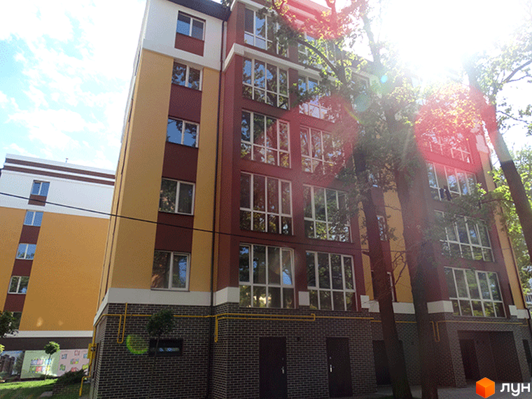 Зовнішній вигляд Житлового комплексу Затишний. Багатоповерховий будинок з великими вікнами та фасадом у жовто-коричневих тонах, поруч дерева.