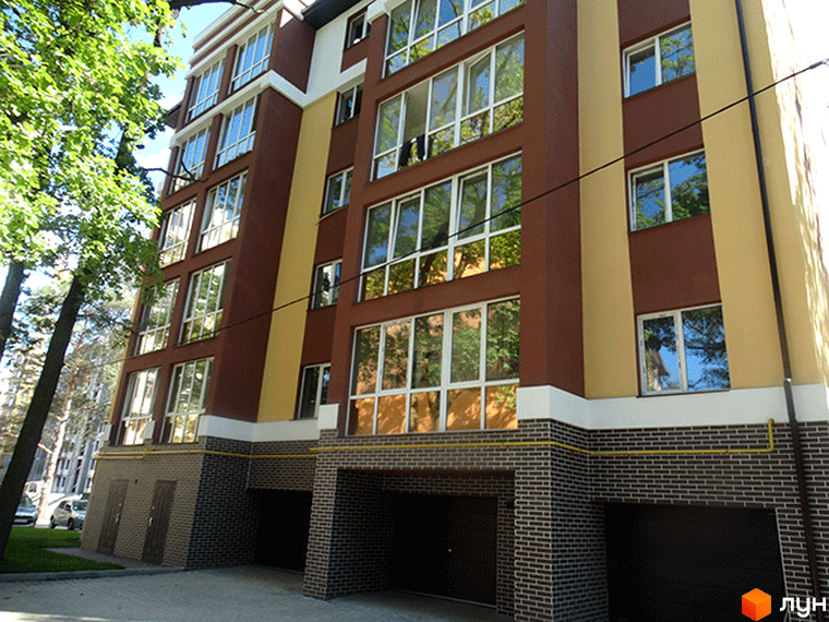 Новобудова Затишний: багатоповерховий будинок з коричнево-бежевим фасадом, великими вікнами. Поруч зелені дерева та асфальтований під'їзд.