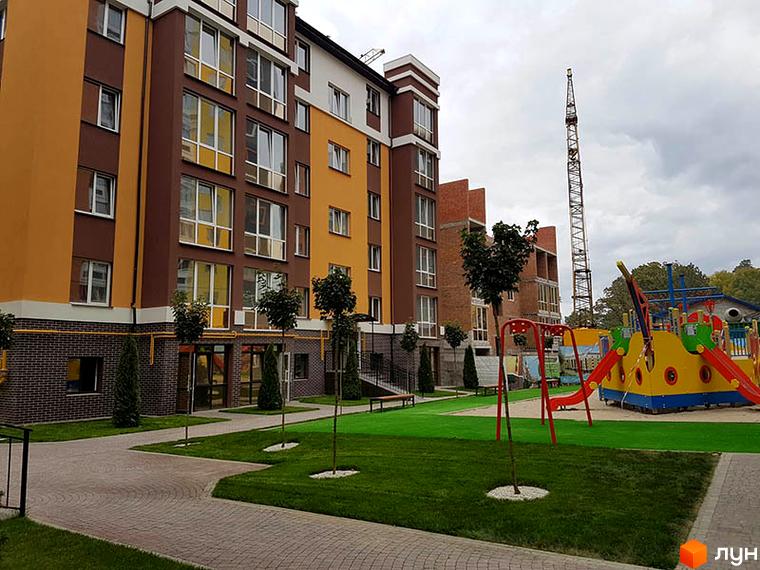 Багатоповерховий будинок Житлового комплексу Затишний з коричнево-жовтим фасадом та великими вікнами. На прибудинковій території зелений газон, доріжки та яскравий дитячий майданчик.