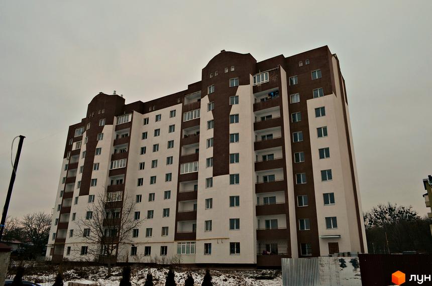 Багатоповерховий будинок з коричнево-білим фасадом, вікнами та балконами. Навколо будівлі видно дерева та сніг.