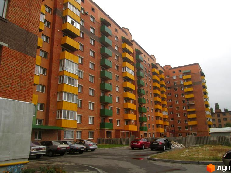 Будинок з цегляним фасадом та яскравими жовтими і зеленими балконами. Прибудинкова територія з автомобілями та зеленими насадженнями.