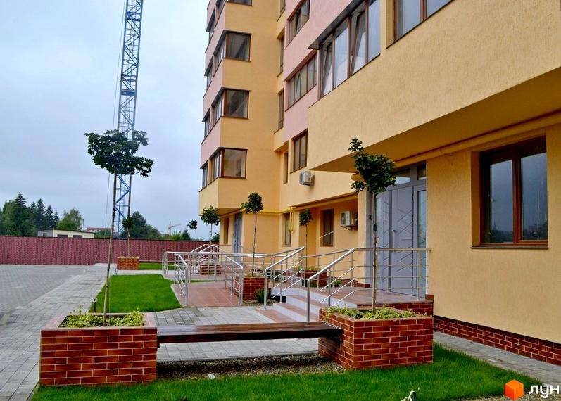 Житловий комплекс Буковинський: світлий фасад, коричневі вікна. Прибудинкова територія з газонами, бруківкою, лавками та пандусами.