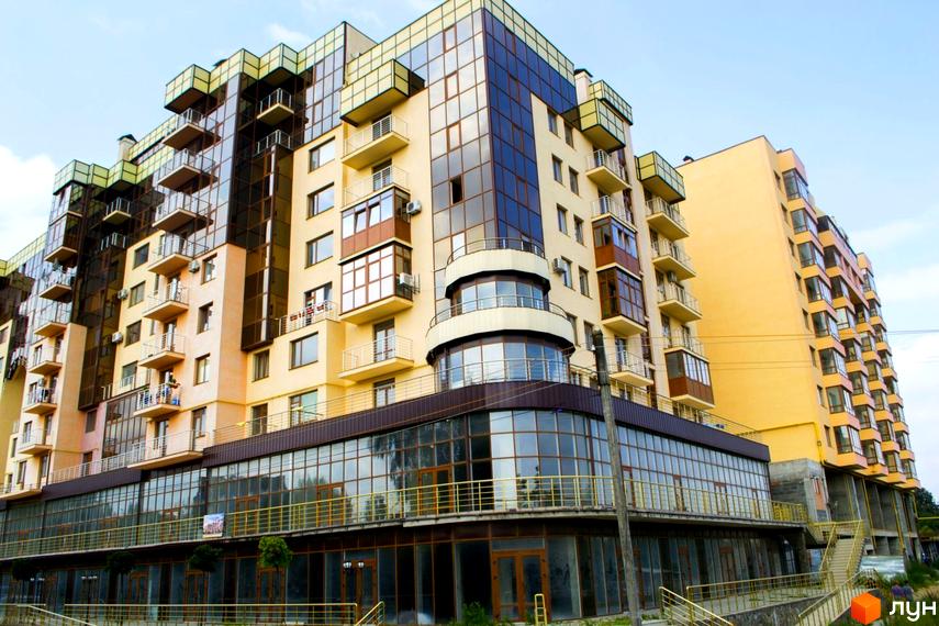 Новобудова Буковинський: багатоповерховий будинок з жовтими стінами та великими скляними секціями. На зображенні видно балкони та комерційні приміщення на першому поверсі.