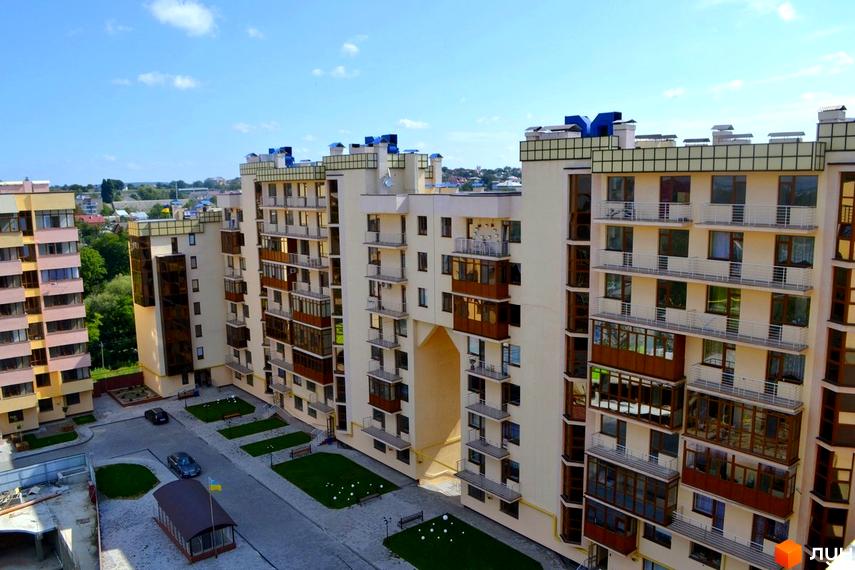 Житловий комплекс Буковинський: багатоповерхові будинки світлого кольору з балконами та арковим проходом, прибудинкова територія з доріжками та газонами.