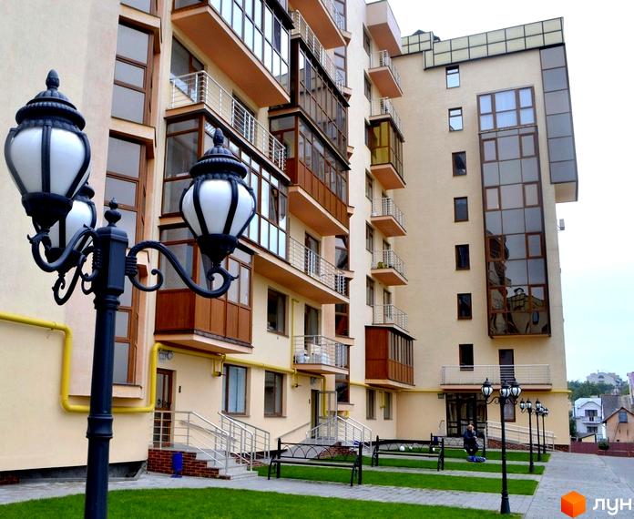 Багатоквартирний будинок Житлового комплексу Буковинський з бежевим фасадом, балконами та великими вікнами. Прибудинкова територія має зелений газон, лавки та вуличні ліхтарі.
