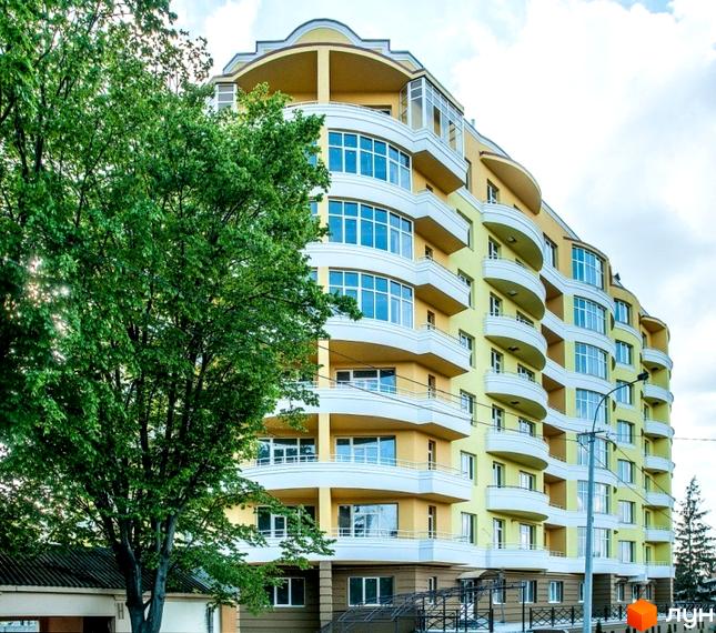 Житловий комплекс світло-жовтого кольору з вигнутими білими балконами та великими вікнами. Перед будинком росте велике зелене дерево.