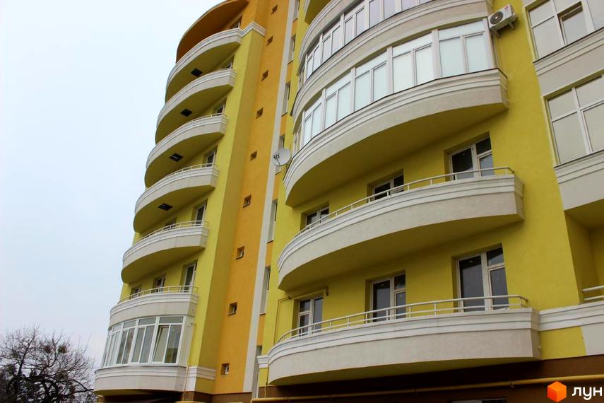 Багатоповерховий житловий будинок жовто-помаранчевого кольору з вигнутими балконами та білими вікнами. Новобудова на вул. Квітки-Основ'яненко, 8б, з прилеглими деревами.