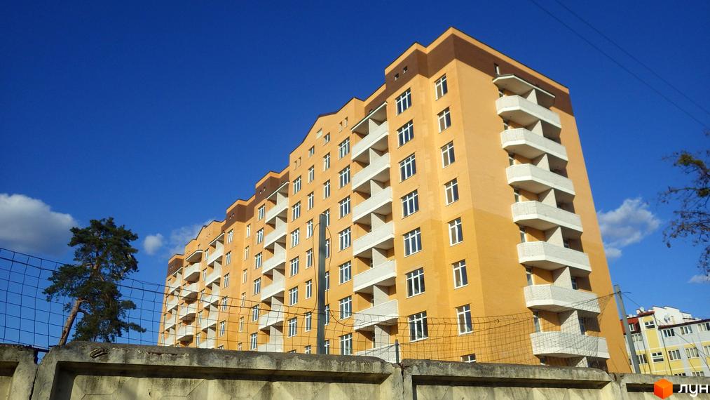 Зовнішній вигляд новобудови ЖК Захисник з жовтою цегляною фасадною частиною та білими балконами на фоні блакитного неба. Навколо новобудови видно паркан та зелене дерево.