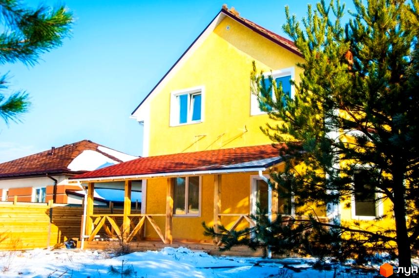 Жовтий двоповерховий будинок з червоним дахом та дерев'яною терасою, розташований у Котеджному містечку HappyTown серед снігу та дерев.