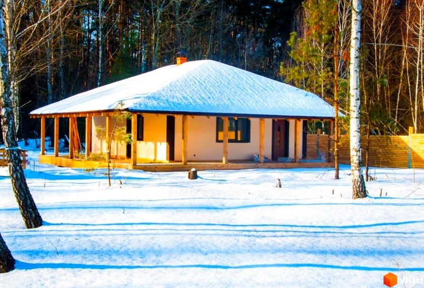 Засніжений одноповерховий будинок з білим фасадом та дерев'яною верандою. Розташований серед лісу в Котеджному містечку HappyTown.