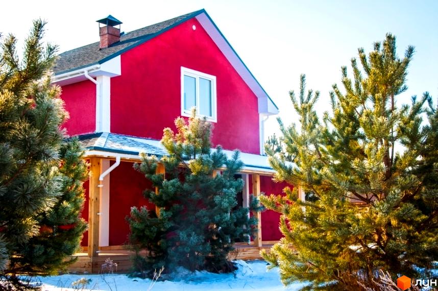 Червоний двоповерховий будинок з білими вікнами та водостоками, оточений зеленими деревами на снігу. Зображення Котеджне містечко HappyTown.