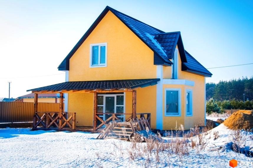 Жовтий двоповерховий будинок з темним дахом та дерев'яною верандою на снігу. Котеджне містечко HappyTown в зимовому пейзажі.