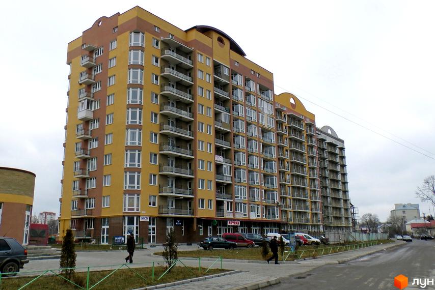 Житловий комплекс Оболоня-парк - багатоповерховий будинок жовто-коричневого кольору з балконами та великою кількістю вікон. Територія навколо будинку з зеленими насадженнями та доріжками.