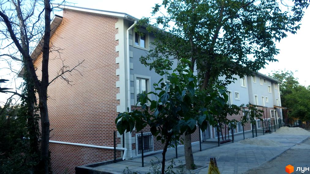Зовнішній вигляд новобудови ЖК Голосіївські сади з цегляним фасадом та сірими елементами. Прибудинкова територія з тротуарною плиткою та деревами.