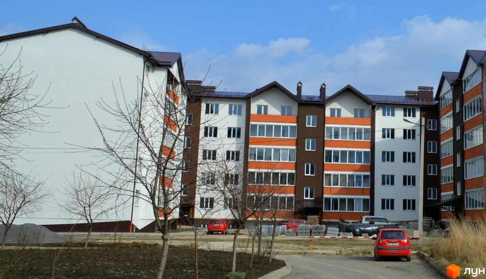 Зовнішній вигляд житлового комплексу Гарантія з білими, коричневими та цегляними фасадами. На прибудинковій території видно дерева та червоний автомобіль.