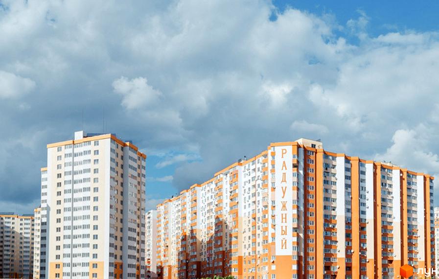 Житловий комплекс Райдужний – це багатоповерхові будинки з білими та помаранчевими фасадами, що розташовані на фоні блакитного неба з хмарами.