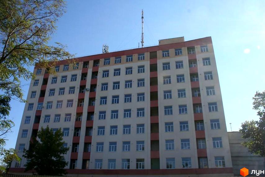 Зовнішній вигляд багатоповерхового будинку ЖК Сонячний Острів з численними вікнами та червоними елементами фасаду. Навколо будівлі видно зелені дерева та синє небо.