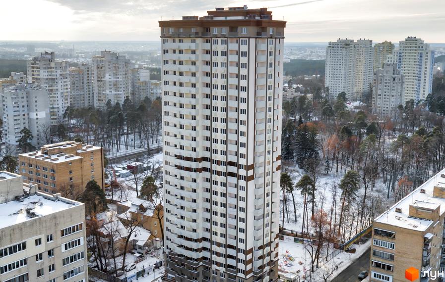 Вид на новобудову ЖК Святобор з фасадом з білої та коричневої плитки, балконами та великою кількістю вікон. Навколо розташовані дерева та інші житлові будинки.