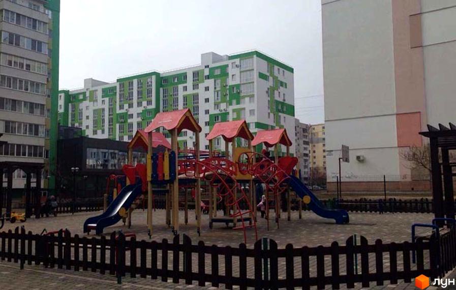 ЖМ Острови: Багатоповерхові будинки з біло-зеленими фасадами та великий дитячий майданчик з яскравими гірками й огорожею.
