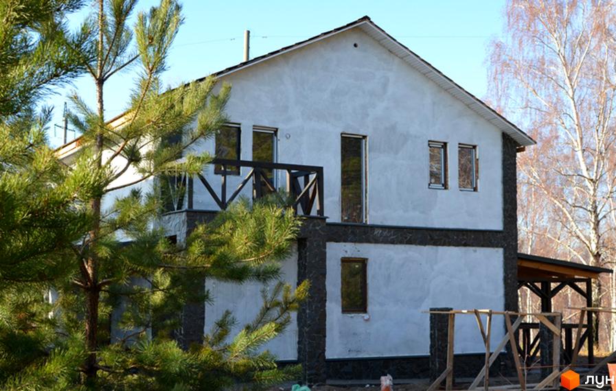Котеджне містечко КМ Смарагдовий: двоповерховий будинок з білою штукатуркою та кам'яним оздобленням цоколя. Навколо будинку зелені сосни та видніються дерева з голим гіллям.