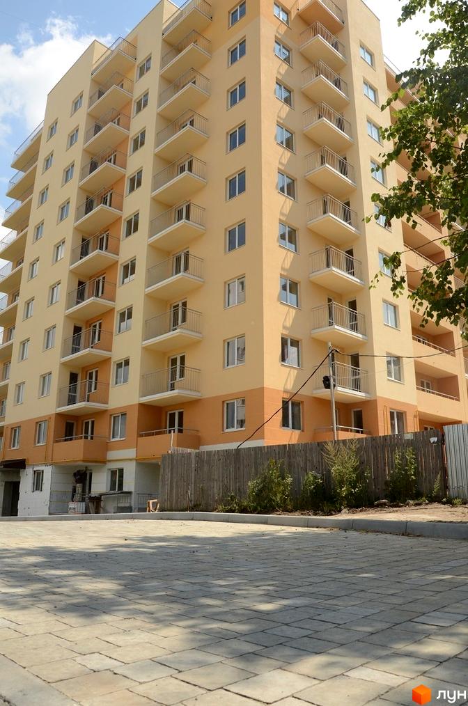 Багатоповерховий житловий будинок світло-жовтого та помаранчевого кольорів з балконами. На передньому плані вимощена бруківка та дерев'яний паркан.