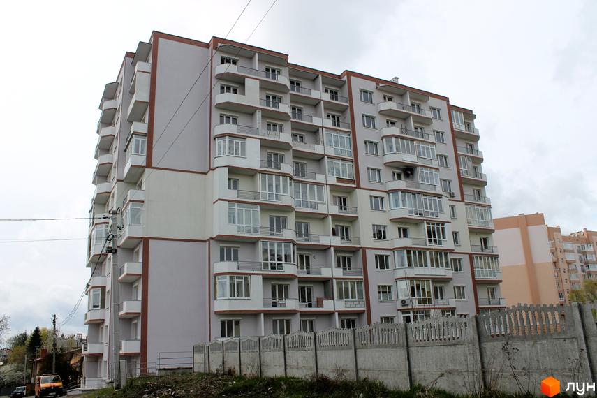 Зовнішній вигляд новобудови на вул. Еммануїла Миська (Врубеля), 15. Багатоповерховий будинок світло-сірого кольору з коричневими елементами та численними балконами. Попереду видно бетонну огорожу та зелень.