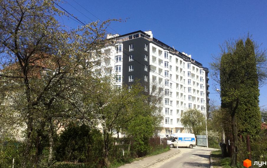 Новобудова ЖК Kaiser з біло-чорним фасадом та численними балконами. Перед будинком ростуть дерева, а дорогою їде білий фургон.