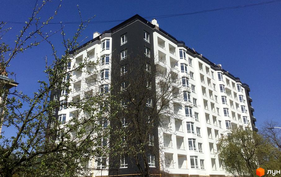 Новобудова ЖК Kaiser з біло-чорним фасадом та балконами на тлі блакитного неба. На передньому плані видно гілки дерев з молодим листям.