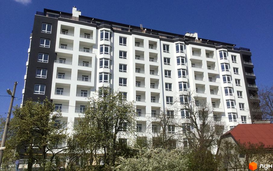 Новобудова ЖК Kaiser з біло-чорним фасадом та численними вікнами. Навколо будівлі видно дерева з квітами та блакитне небо.