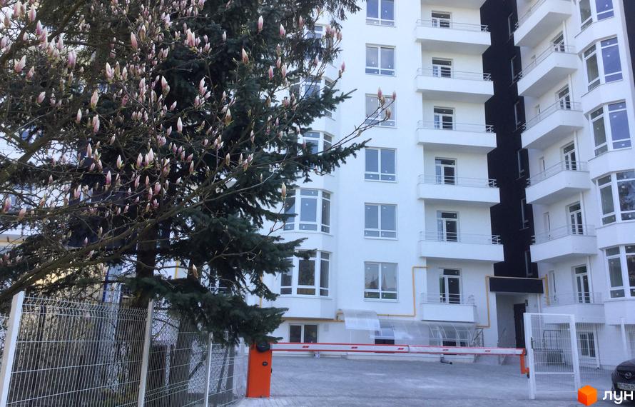 Житловий комплекс Kaiser – біла багатоповерхівка з балконами та вікнами. На передньому плані дерево з бутонами та огорожа.