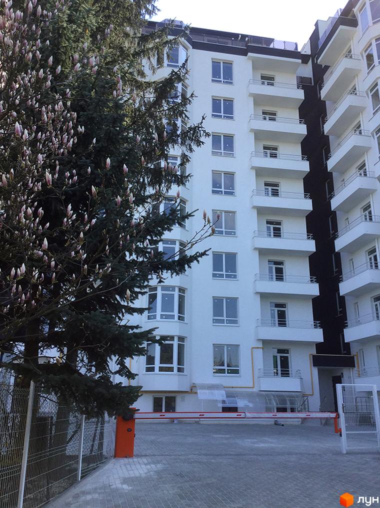 Житловий комплекс Kaiser: біла багатоповерхівка з балконами та вікнами, поруч зелені дерева та паркан з автоматичним шлагбаумом.