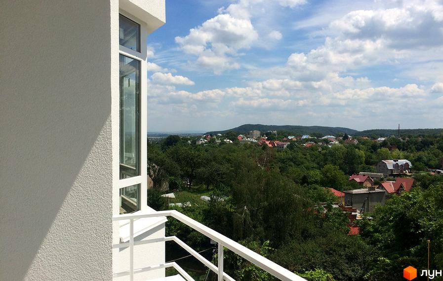 Вид з вікна житлового комплексу Kaiser на зелені пагорби з будиночками. Сучасний фасад будівлі з великими вікнами та балконом.