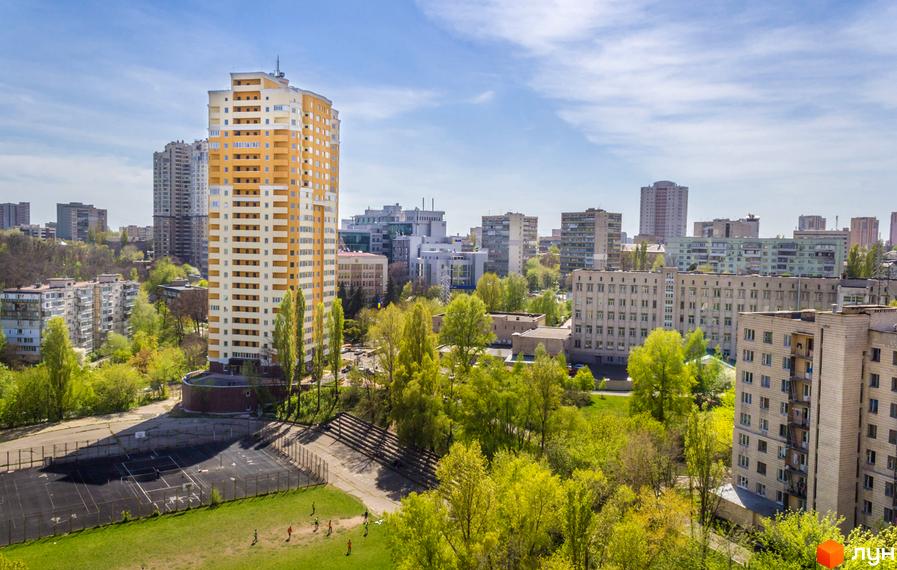 Висотна будівля ЖК Solo Park з жовто-білими фасадами на тлі міської забудови. На передньому плані зелена зона з деревами та спортивний майданчик.