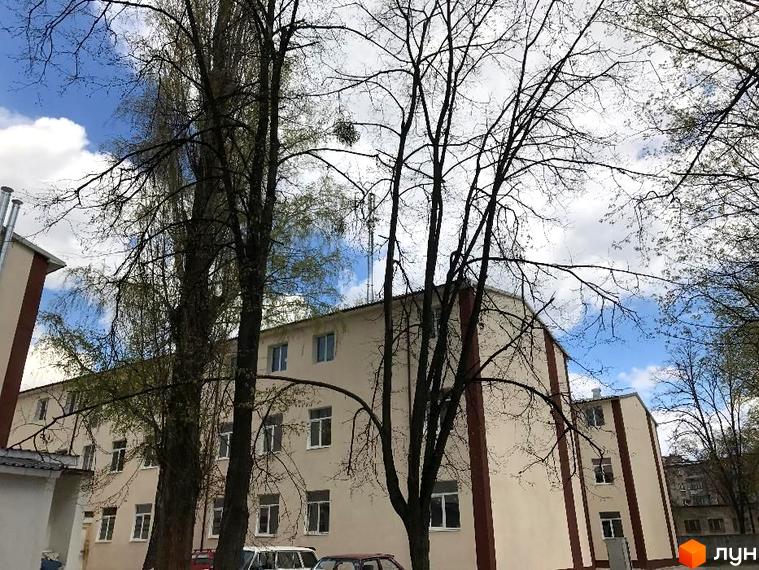 Зовнішній вигляд житлового комплексу Садове місто з фасадом світло-бежевого кольору та червоними вертикальними смугами. Навколо будинку ростуть дерева з гілками без листя.