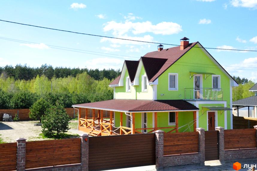 Котеджне містечко HappyTown: Салатовий двоповерховий будинок з коричневим дахом, верандою та балконом. Навколо будинку дерев'яний паркан та зелені насадження.