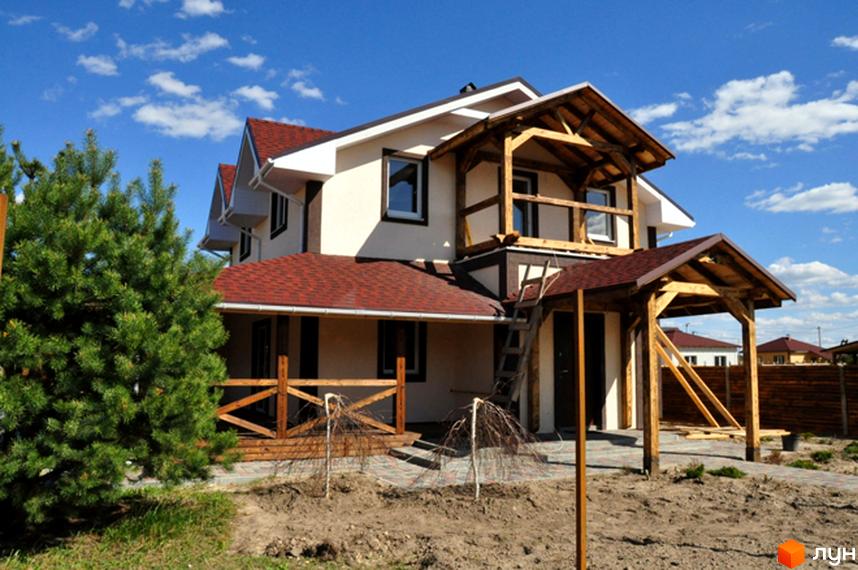 Двоповерховий будинок Котеджного містечка HappyTown з червоним дахом та дерев'яними конструкціями. На ділянці – сосна, молоді дерева та бруківка.