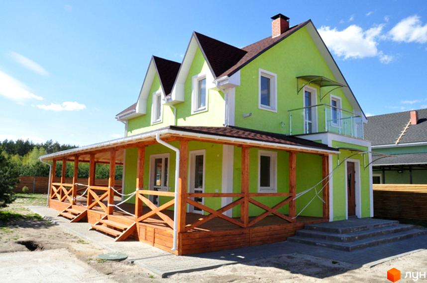 Двоповерховий будинок салатового кольору з коричневим дахом та просторою дерев'яною терасою. Територія Котеджного містечка HappyTown.