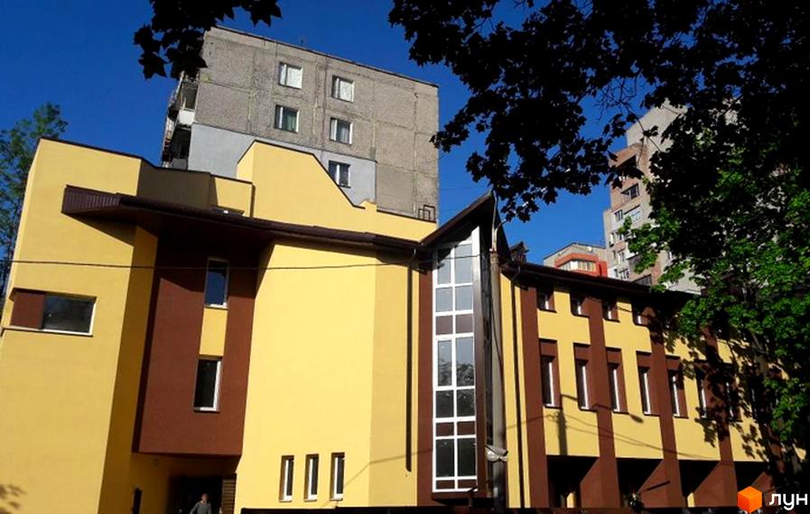 Клубний будинок Княжий: жовто-коричнева фасадна частина будівлі з великими вікнами та сусідній сірий багатоповерховий будинок на фоні блакитного неба.
