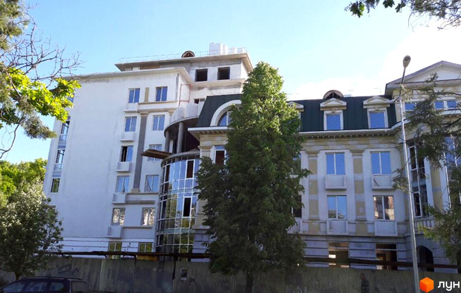 Зовнішній вигляд новобудови ЖК Парковий: біла багатоповерхова будівля з скляним еркером та класичною архітектурою сусідньої будівлі. Навколо зелені дерева та блакитне небо.